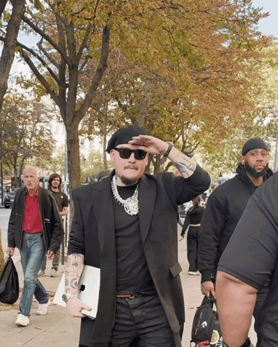 A Man Wearing Sunglasses And A Black Beret Is Walking Down A Sidewalk GIF