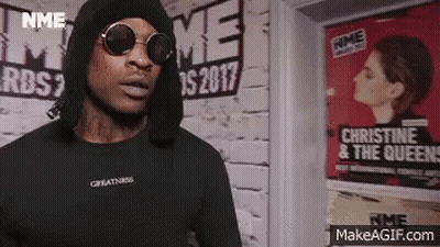 A Man Wearing Sunglasses And A Black Hoodie Is Standing In Front Of A Wall . GIF