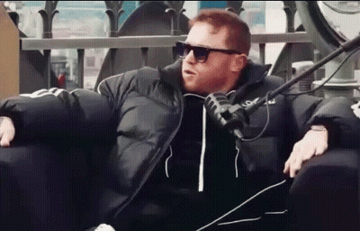 A Man Wearing Sunglasses And A Black Jacket Is Sitting In A Chair With A Microphone . GIF
