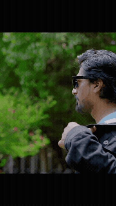 A Man Wearing Sunglasses And A Black Jacket Stands In Front Of Trees GIF