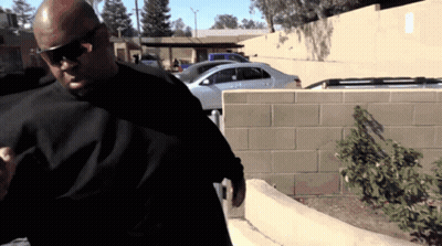 A Man Wearing Sunglasses And A Black Shirt Stands In Front Of A Parking Lot GIF