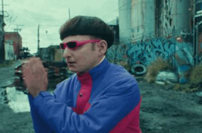 A Man Wearing Sunglasses And A Blue And Pink Jacket Is Standing In Front Of Graffiti GIF
