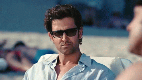 A Man Wearing Sunglasses And A Blue Shirt Is Sitting On A Beach GIF