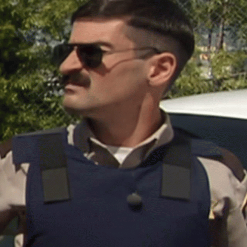 A Man Wearing Sunglasses And A Blue Vest Is Standing In Front Of A Car GIF