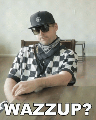 A Man Wearing Sunglasses And A Checkered Shirt Is Sitting At A Table With The Words Wazzup On The Table GIF