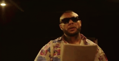 A Man Wearing Sunglasses And A Colorful Shirt Is Standing In A Dark Room Holding A Piece Of Paper . GIF