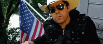 A Man Wearing Sunglasses And A Cowboy Hat Stands In Front Of An American Flag GIF