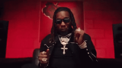 A Man Wearing Sunglasses And A Cross Necklace Is Giving A Thumbs Up In Front Of A Red Wall . GIF