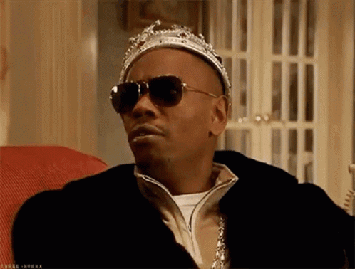 A Man Wearing Sunglasses And A Crown Is Sitting In A Chair . GIF