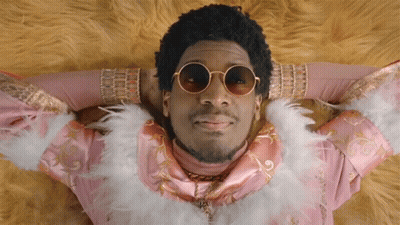 A Man Wearing Sunglasses And A Feather Boa Is Laying Down GIF