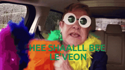 A Man Wearing Sunglasses And A Feather Boa Says " Hee Shaall Bee Le Veon " GIF