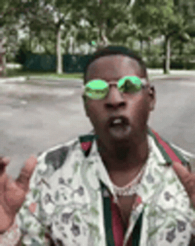 A Man Wearing Sunglasses And A Floral Shirt Is Standing In Front Of A Street . GIF