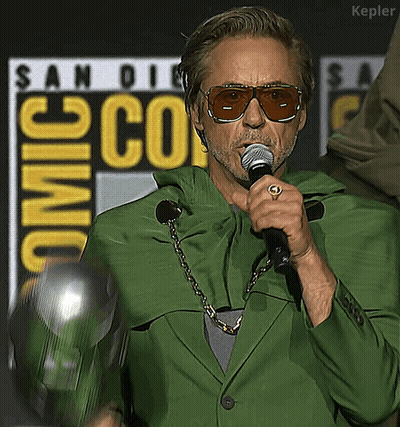 A Man Wearing Sunglasses And A Green Jacket Is Holding A Microphone In Front Of A San Diego Comic Con Sign GIF