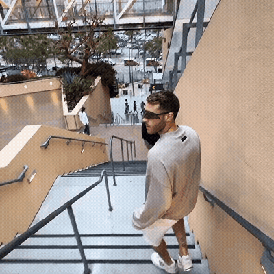A Man Wearing Sunglasses And A Grey Sweatshirt Is Walking Down Stairs GIF