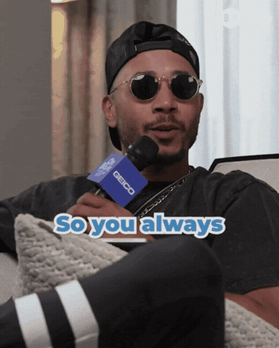 A Man Wearing Sunglasses And A Hat Is Holding A Microphone That Says Geico On It GIF