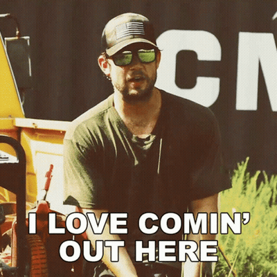 A Man Wearing Sunglasses And A Hat Is Sitting In Front Of A Sign That Says " I Love Comin ' Out Here " GIF
