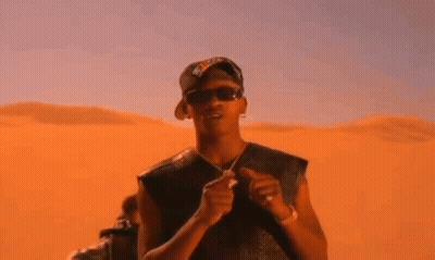 A Man Wearing Sunglasses And A Hat Is Standing In Front Of A Sand Dune . GIF