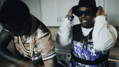 A Man Wearing Sunglasses And A Hat Is Standing Next To Another Man Wearing A Purple Vest GIF