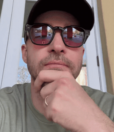 A Man Wearing Sunglasses And A Hat Looks At The Camera With His Hand On His Chin GIF