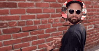 A Man Wearing Sunglasses And A Hat Stands In Front Of A Red Brick Wall GIF