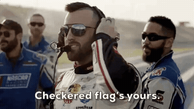 A Man Wearing Sunglasses And A Headset Is Standing Next To A Group Of Race Car Drivers On A Race Track . GIF