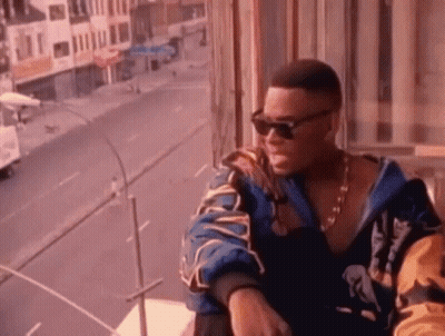 A Man Wearing Sunglasses And A Jacket Is Sitting On A Balcony Overlooking A Street . GIF