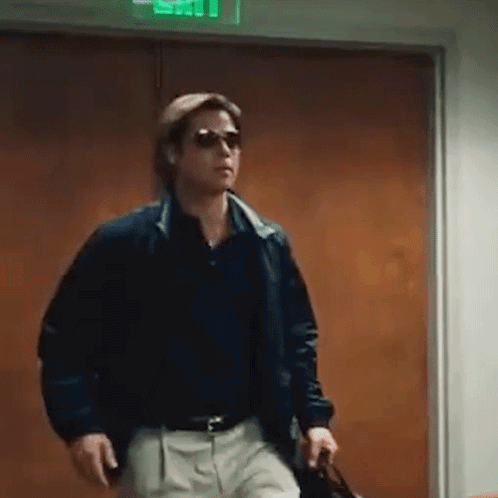 A Man Wearing Sunglasses And A Jacket Is Walking Through A Doorway Holding A Briefcase . GIF