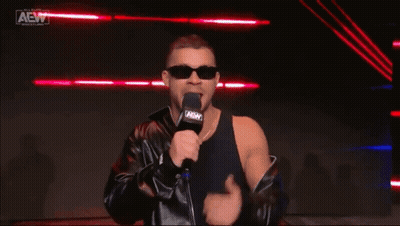 A Man Wearing Sunglasses And A Leather Jacket Is Holding A Microphone And Covering His Eyes . GIF