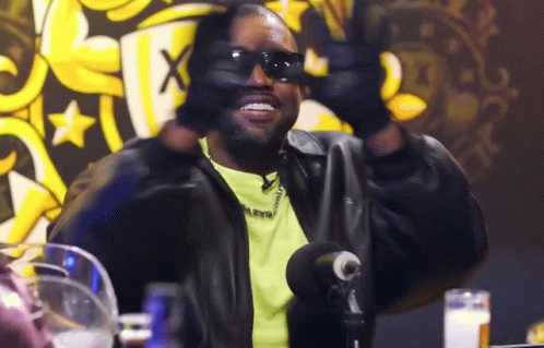 A Man Wearing Sunglasses And A Leather Jacket Is Sitting At A Table With A Microphone . GIF