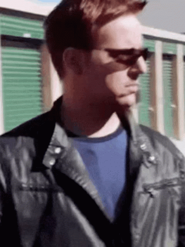 A Man Wearing Sunglasses And A Leather Jacket Stands In Front Of A Green Building . GIF