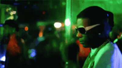 A Man Wearing Sunglasses And A Necklace Is Standing In A Crowded Room . GIF