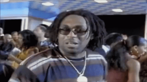 A Man Wearing Sunglasses And A Necklace Is Standing In A Crowded Room . GIF
