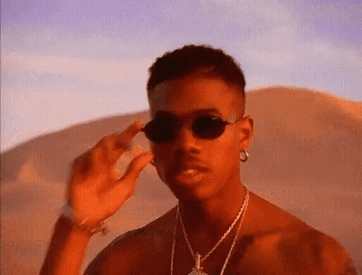 A Man Wearing Sunglasses And A Necklace Is Standing In The Desert . GIF