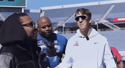 A Man Wearing Sunglasses And A Ny Giants Jersey Talks To Another Man GIF