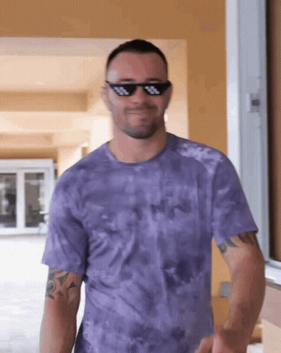 A Man Wearing Sunglasses And A Purple Tie Dye Shirt Is Smiling GIF