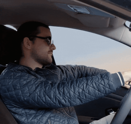 A Man Wearing Sunglasses And A Quilted Jacket Drives A Car GIF