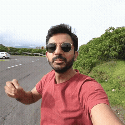 A Man Wearing Sunglasses And A Red Shirt Is Standing On The Side Of A Road GIF
