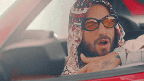 A Man Wearing Sunglasses And A Scarf Is Driving A Car GIF