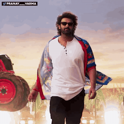 A Man Wearing Sunglasses And A Scarf Is Walking In Front Of A Red Tractor With The Words Pranay Varma Above Him GIF