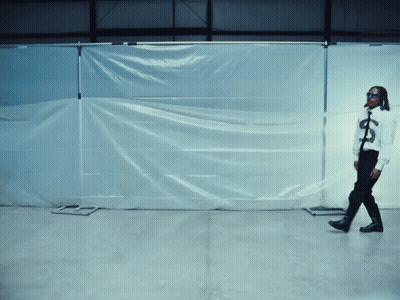 A Man Wearing Sunglasses And A Shirt With A Dollar Sign On It Stands In Front Of A White Tarp GIF