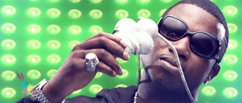 A Man Wearing Sunglasses And A Skull Ring Is Holding A Microphone In Front Of A Green Background GIF