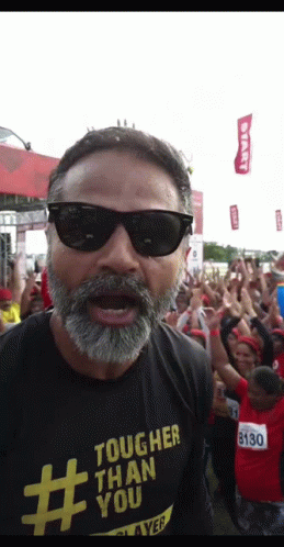 A Man Wearing Sunglasses And A Tougher Than You Shirt Stands In Front Of A Crowd GIF