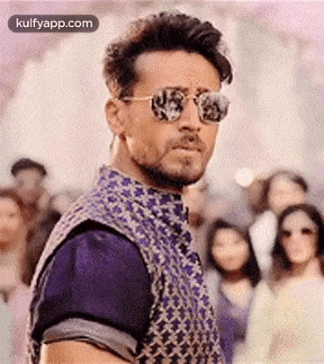 A Man Wearing Sunglasses And A Vest Is Standing In Front Of A Crowd Of People . GIF