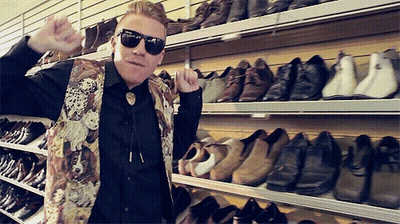 A Man Wearing Sunglasses And A Vest With A Dog On It Stands In Front Of A Wall Of Shoes GIF