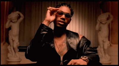A Man Wearing Sunglasses And A Watch Is Sitting In Front Of A Statue Of A Woman GIF