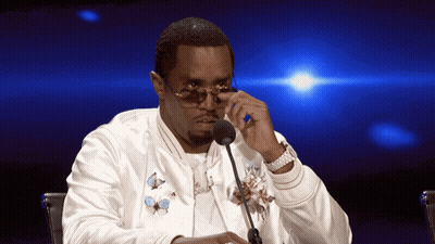 A Man Wearing Sunglasses And A White Jacket Is Sitting At A Table With A Microphone GIF