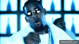 A Man Wearing Sunglasses And A White Shirt Is Singing Into A Microphone In Front Of A Blue Background . GIF