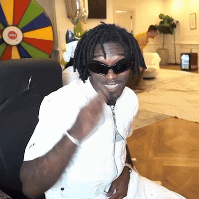 A Man Wearing Sunglasses And A White Shirt Is Sitting In Front Of A Wheel Of Fortune GIF