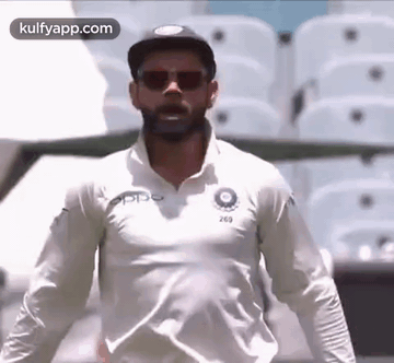 A Man Wearing Sunglasses And A White Shirt Is Standing In Front Of A Stadium . GIF