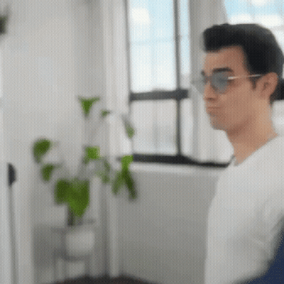 A Man Wearing Sunglasses And A White Shirt Is Standing In Front Of A Window In A Room . GIF